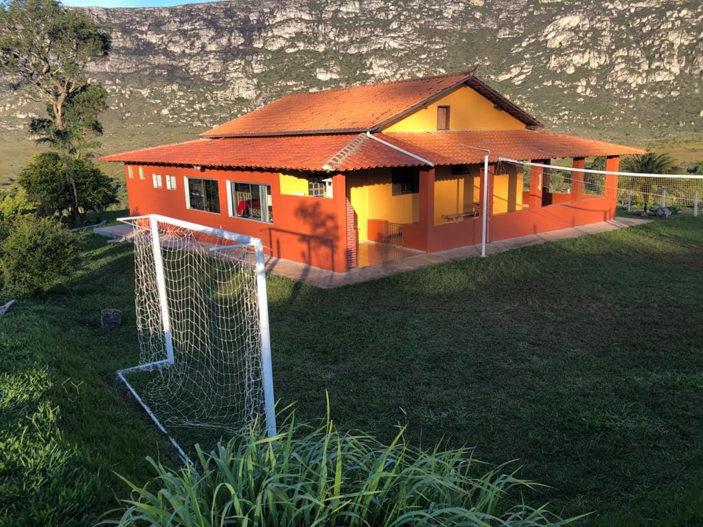 A vibrant yellow house with a red roof is nestled in a verdant landscape, featuring expansive windows that provide views of the surrounding mountains. A grassy area suitable for sports is visible in front of the house, complete with a soccer goal and open space for various outdoor activities.