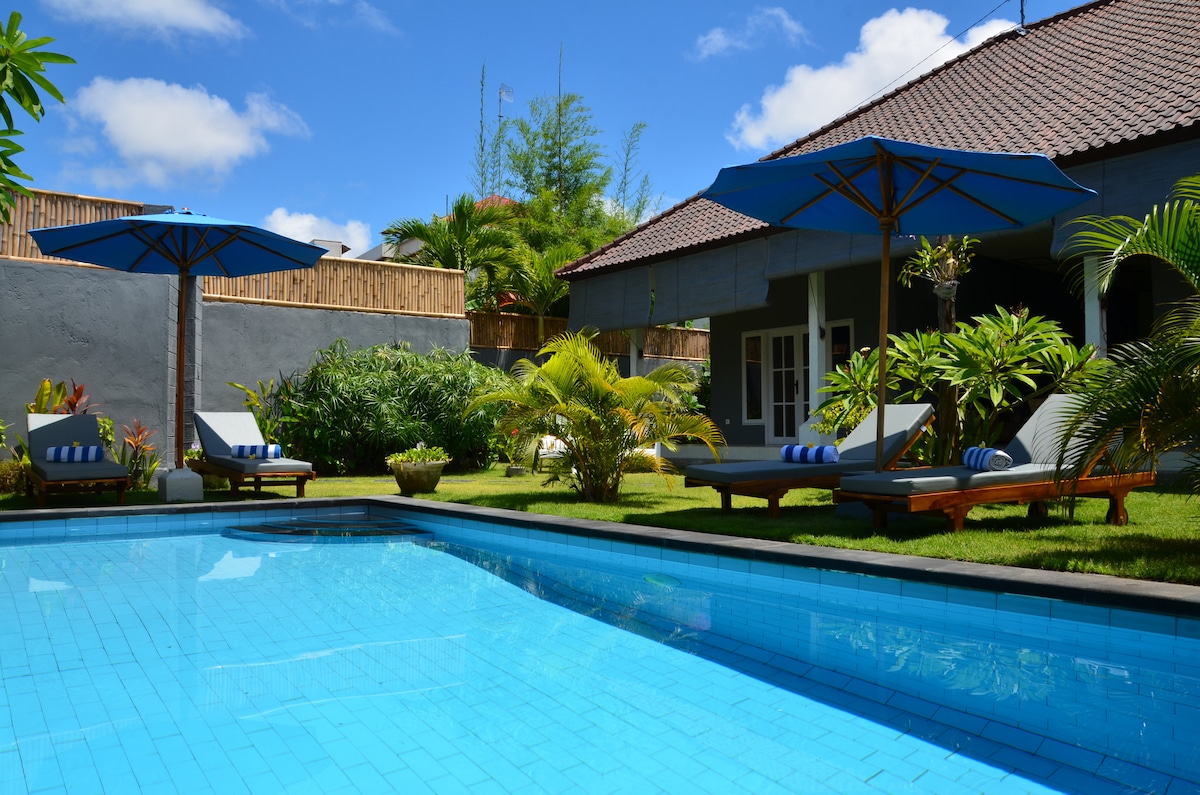 A refreshing pool is surrounded by lush greenery and well-maintained grass. Lounge chairs with blue umbrellas offer shaded seating. The backdrop features a single-story building with large windows, contributing to a serene outdoor space.