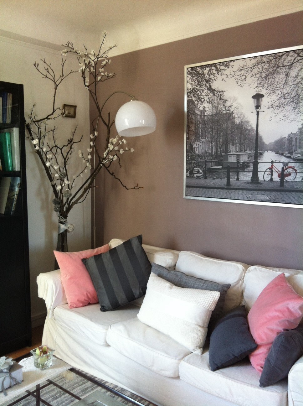 A living room corner is highlighted by a white sofa adorned with pillows in shades of pink and gray. A large black-and-white photograph hangs on the wall, depicting a scenic street. A decorative branch with artificial flowers adds a touch of nature to the space.