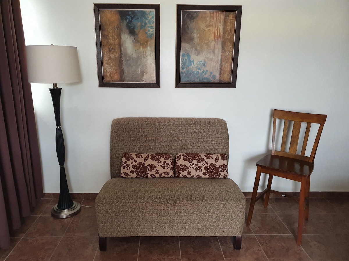 A small seating area features a patterned sofa with two decorative cushions. Adjacent to the sofa, a wooden chair is positioned nearby. The space is complemented by a floor lamp and two framed art pieces that hang on the wall.