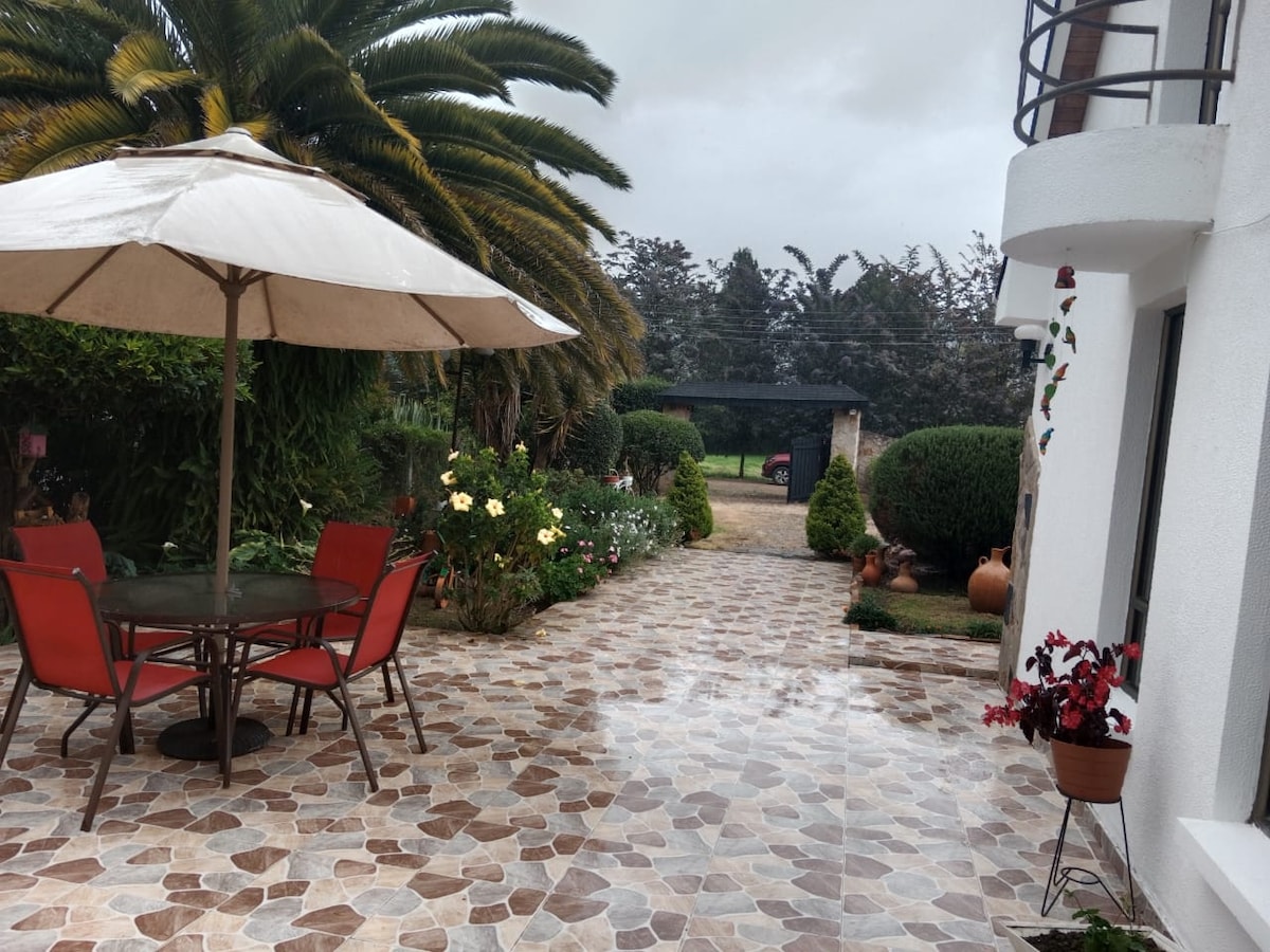 An outdoor seating area features a round table with four chairs under a large umbrella. Lush greenery and colorful flowers surround the paved patio, while a shaded area is visible in the background, providing additional space for relaxation.