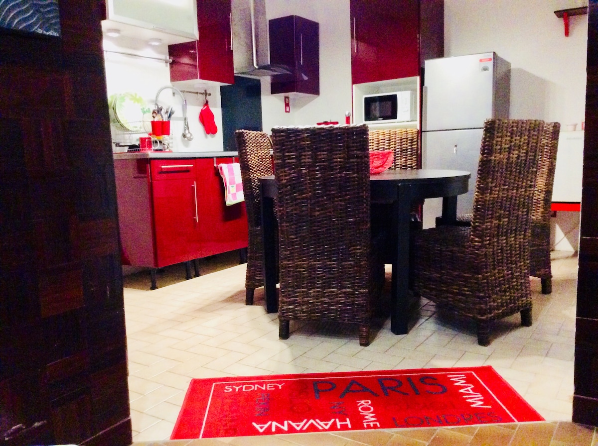 The kitchen area features red cabinetry and a dining table surrounded by six woven chairs. A modern refrigerator and a microwave are visible, and a decorative rug with city names adds a touch of interest to the spacious tiled floor.