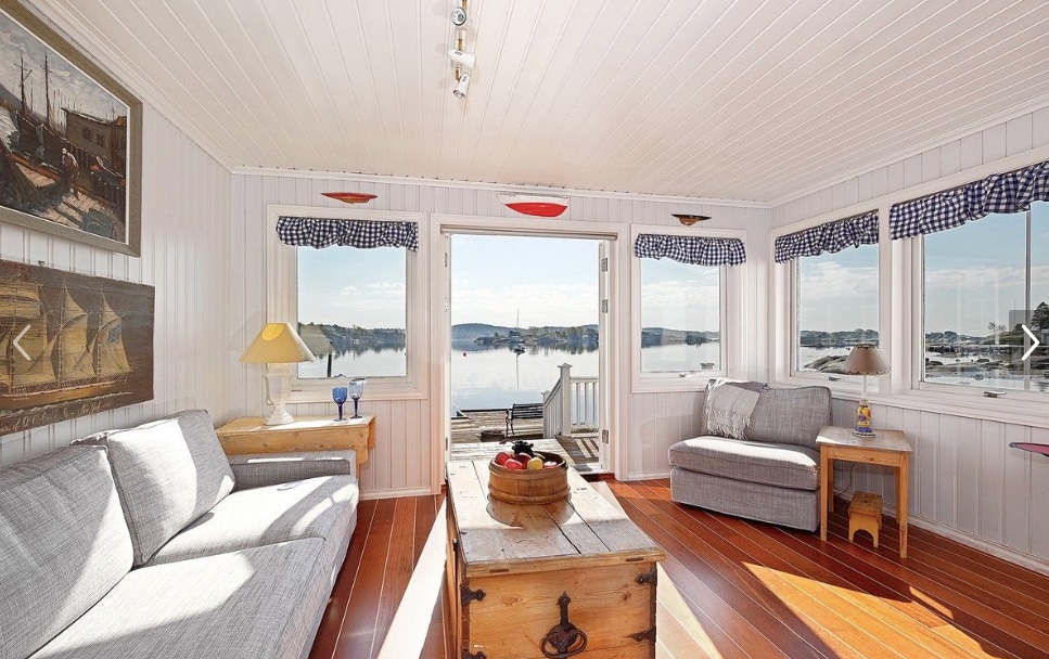 A light-filled glass enclosed porch features two comfortable seating areas, with a coffee table positioned between them. Large windows provide views of the water and shoreline, while walls are adorned with nautical-themed artwork and traditional window treatments in a checkered pattern.
