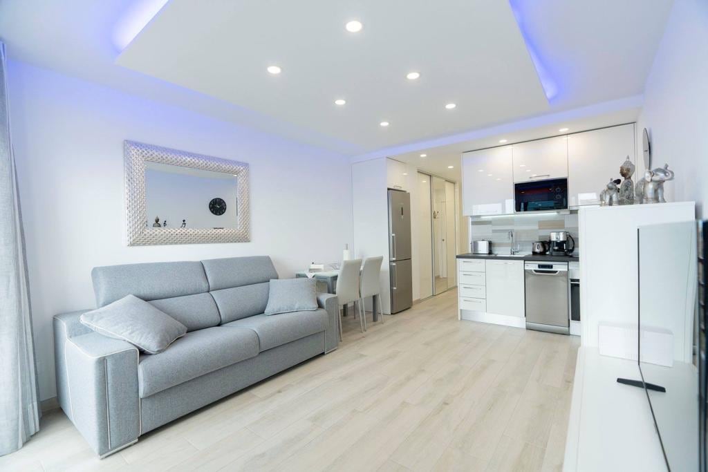A modern living area features a comfortable gray sofa, complemented by soft pillows. A minimalist dining table with white chairs is visible in the background. The kitchenette, equipped with contemporary appliances, is accented by bright lighting, contributing to the airy atmosphere of the space.