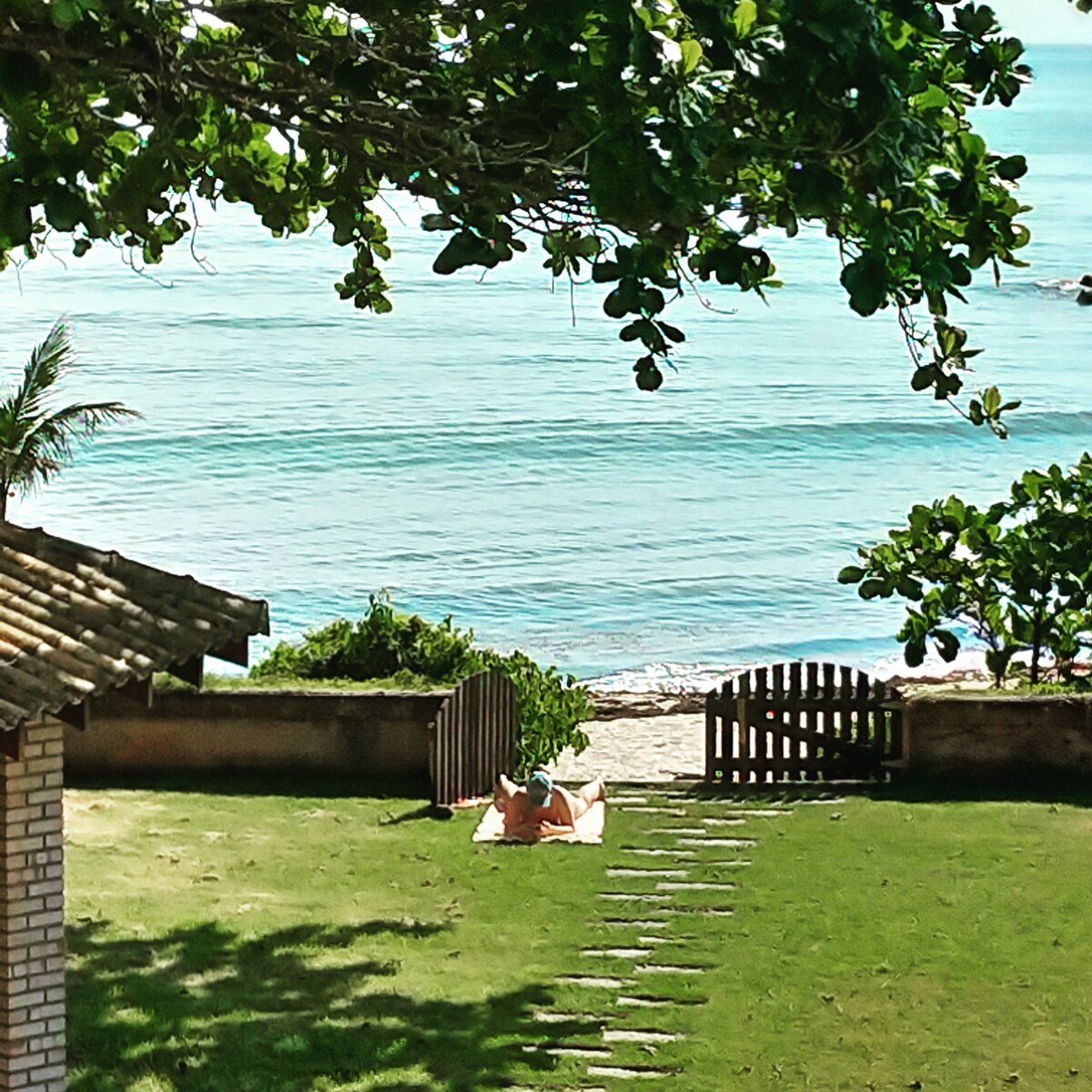 A peaceful outdoor area features a well-maintained lawn leading to a sandy beach. Two chairs are positioned near the water, framed by lush greenery and trees. The ocean is visible in the background, with gentle waves lapping at the shore.