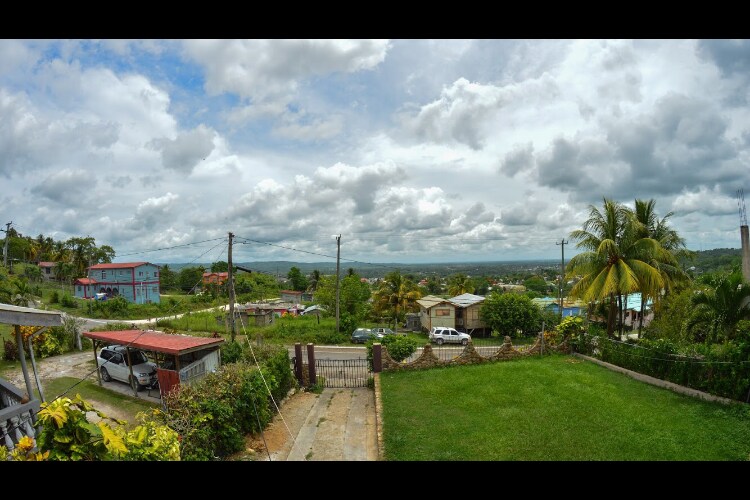 A panoramic view showcases a vibrant landscape with green hills and scattered residential buildings. The sky is filled with fluffy clouds, and patches of blue are visible. A small lawn extends in the foreground, bordered by a stone wall and a driveway leading to parked vehicles.