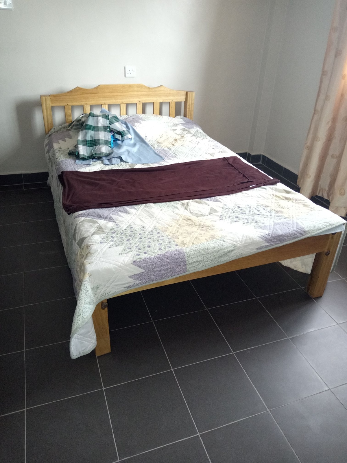 A spacious bed is positioned in the corner of the room, featuring a patterned bedspread. A green and white checkered shirt and a maroon cloth are casually placed on the bed. Natural light filters through a nearby window, illuminating the tiled flooring.