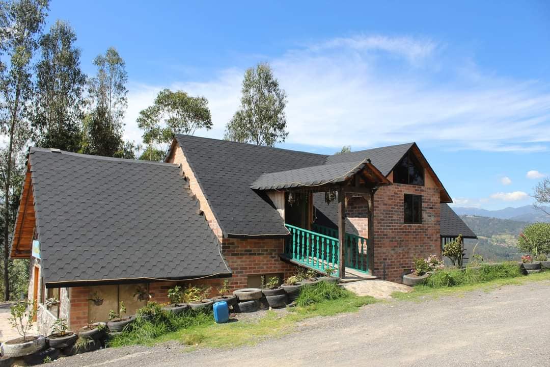 The exterior of a rustic house is framed by rolling hills and tall trees. Brick walls and a slate roof complement the natural surroundings. A pathway leads to a welcoming entrance adorned with a green railing, inviting guests to explore the tranquil retreat.