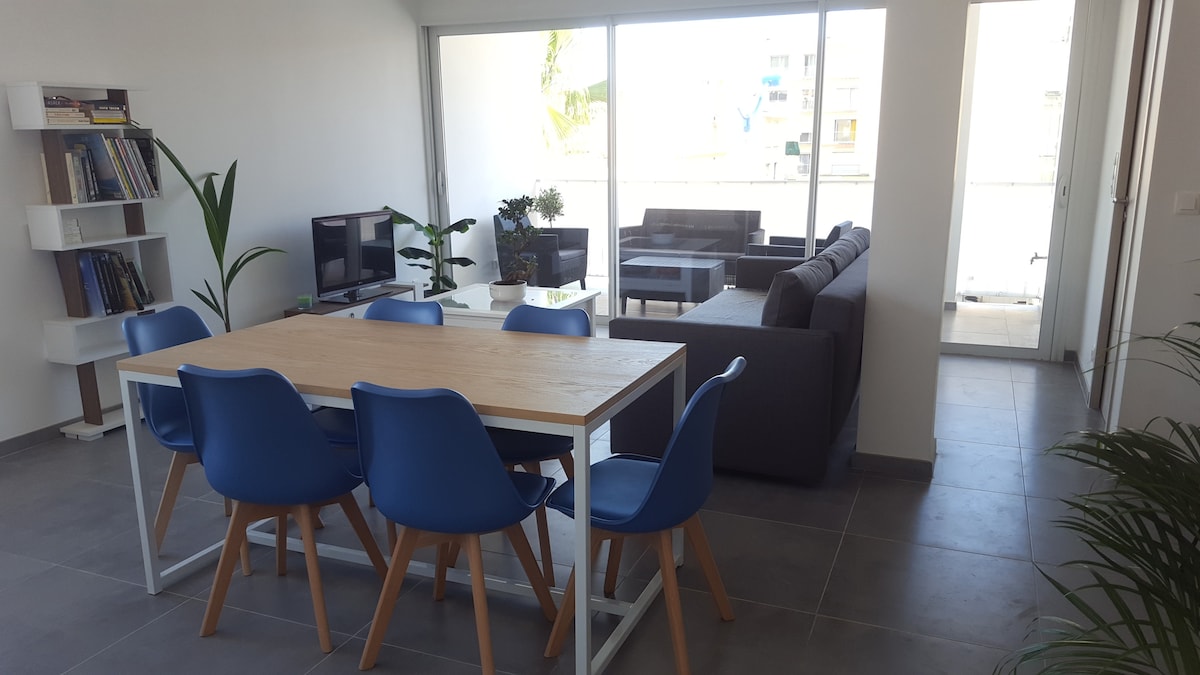 A modern living space is shown, featuring a spacious dining table surrounded by blue chairs. A comfortable seating area with dark sofas is visible, along with plants that add a touch of greenery. Large glass doors allow ample natural light, enhancing the openness.