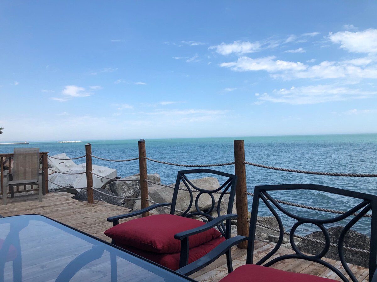 A view of the lake from a wooden deck, showcasing clear blue waters and a bright sky. Metal chairs with comfortable red cushions are positioned around a glass table, enhancing the outdoor seating area. Natural stone and wooden posts define the perimeter of the deck.