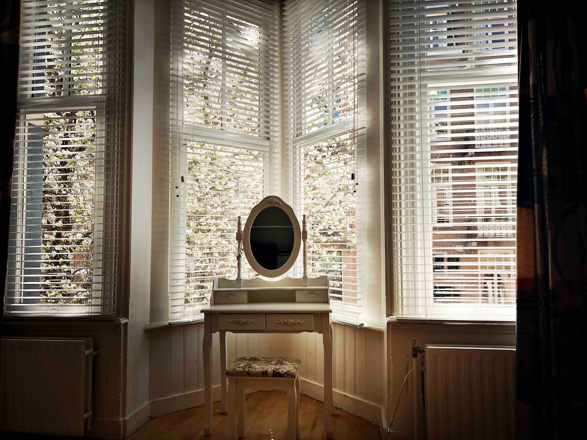 A charming vanity table with a round mirror is positioned in front of large bay windows. The windows feature white blinds, allowing soft light to filter into the room. A patterned stool adds a touch of design, complementing the natural wood flooring.