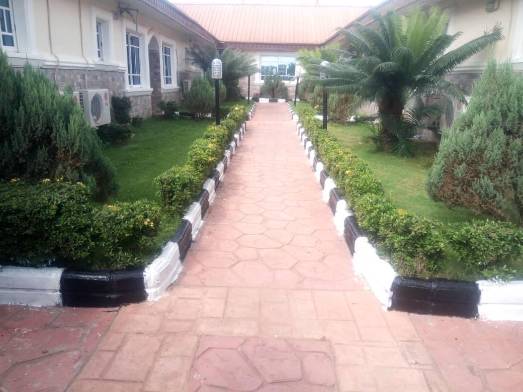 A well-maintained path of textured tiles leads through a vibrant garden, bordered by neatly trimmed hedges and decorative plants. Colorful landscaping elements enhance the ambience, with light reflecting off the path's edges painted in black and white, guiding the way towards a welcoming entrance.