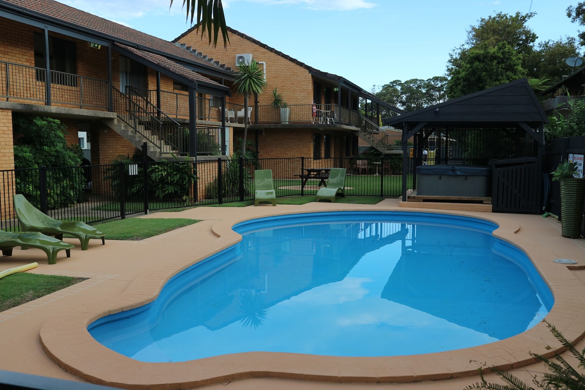 A shared outdoor pool is surrounded by a spacious, landscaped garden. Sun loungers are positioned nearby, and a gazebo provides shaded seating. The two-story apartment complex is visible in the background, showcasing balconies overlooking the pool area.