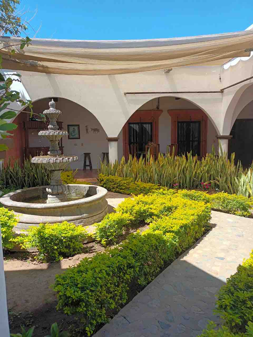 A central courtyard is showcased with a stone fountain surrounded by greenery and neatly manicured hedges. Arched architecture adds to the aesthetic, while shaded areas provide relief from the sun. The natural light enhances the inviting atmosphere of the outdoor space.