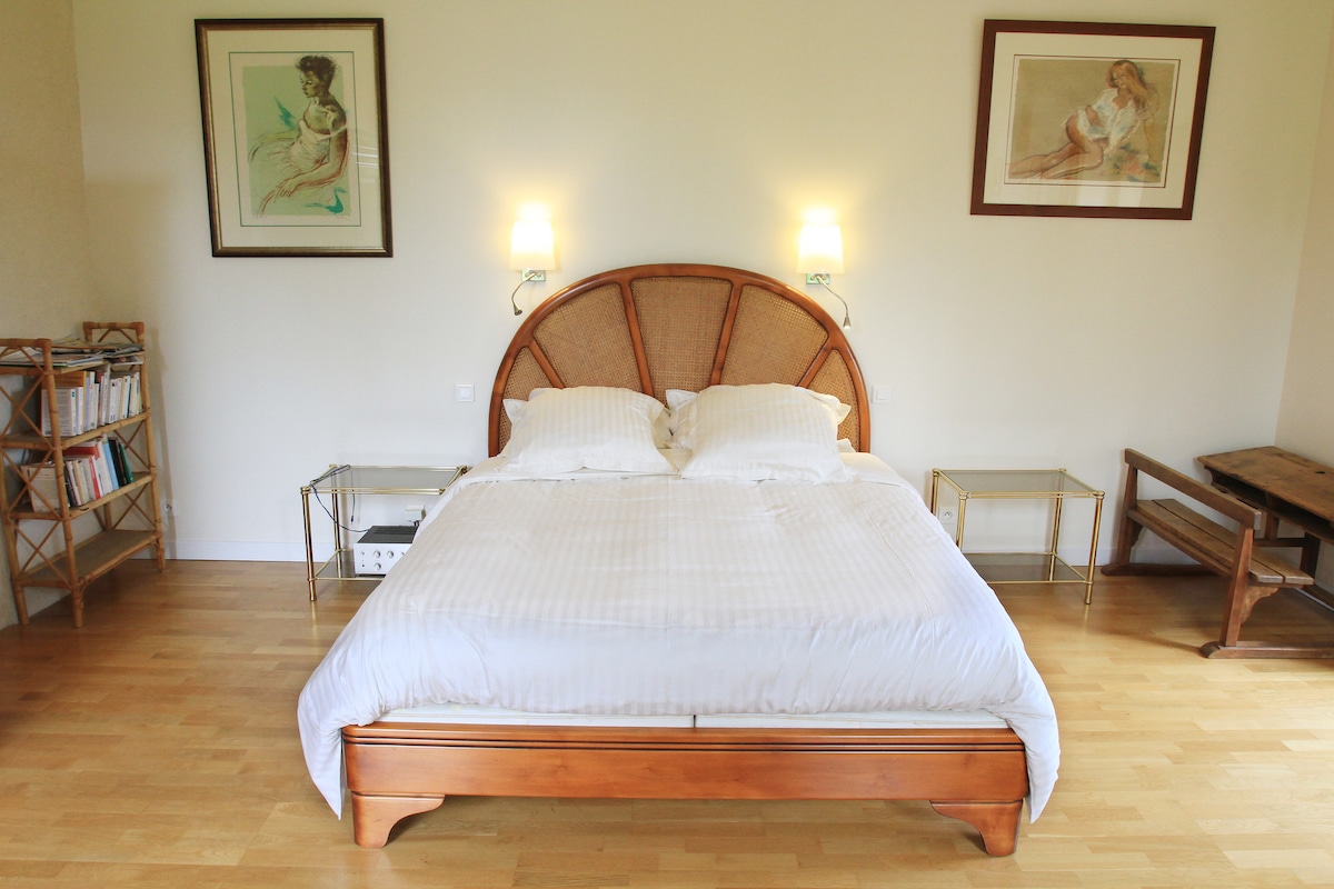 A spacious bedroom is furnished with a wooden bed set against a light wall. Two bedside tables are positioned beside the bed, and artwork hangs above, adding a touch of elegance. A wooden desk and a small bookshelf are arranged to one side, enhancing the room's functionality.