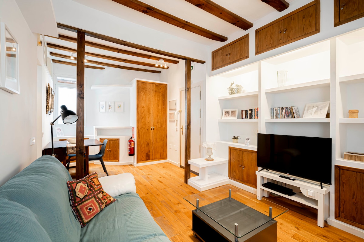 The living room features wooden beams and light-colored walls, creating a bright and airy environment. Shelves display decorative items and books. A comfortable sofa with cushions is positioned near a stylish coffee table and a flat-screen TV, enhancing the functionality of the space.