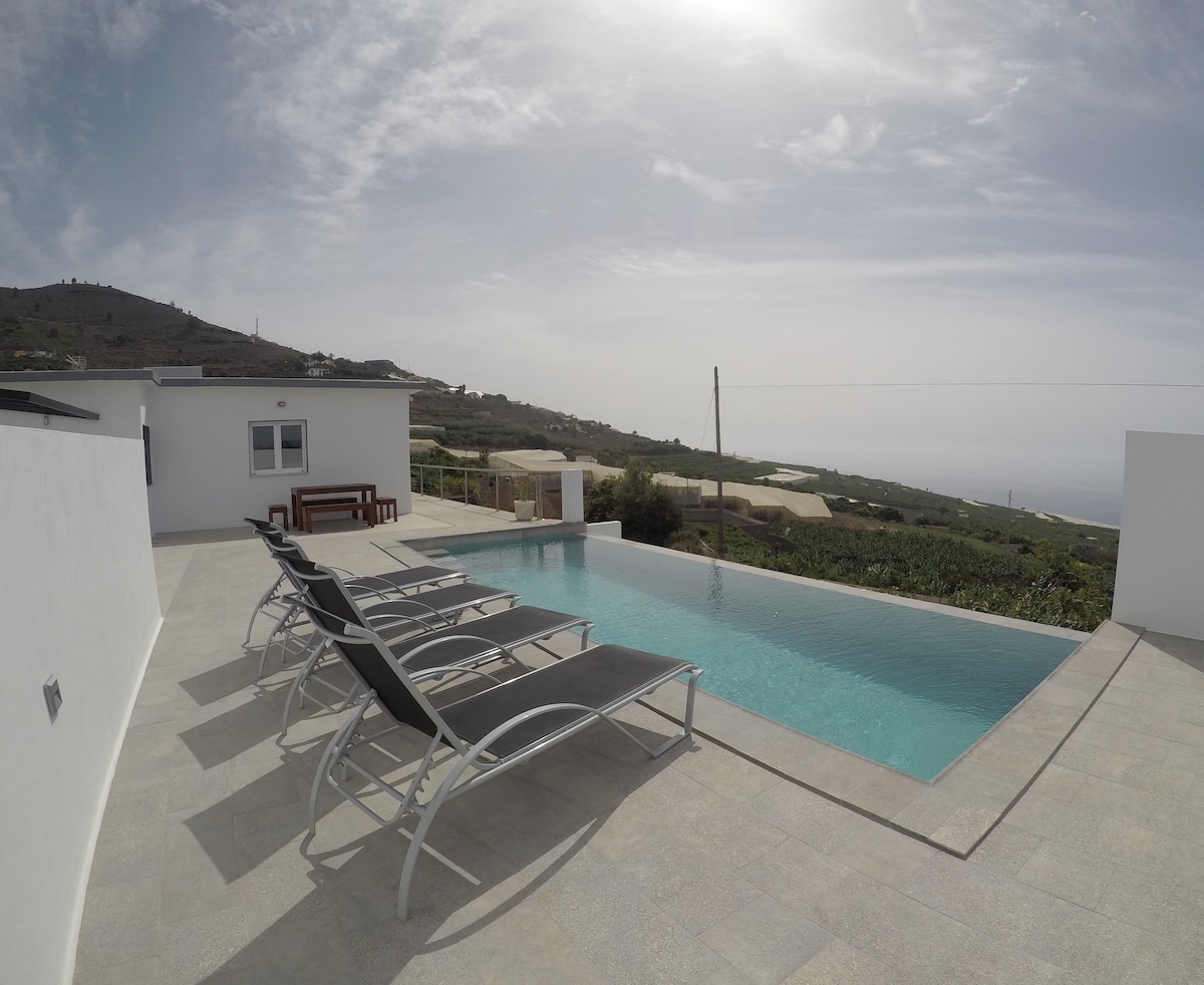 A modern outdoor space features a pool surrounded by lounge chairs, providing a serene area for relaxation. The pool reflects the sky, and the surrounding landscape displays soft hills and greenery. A shaded seating area is visible in the background, enhancing the tranquil setting.
