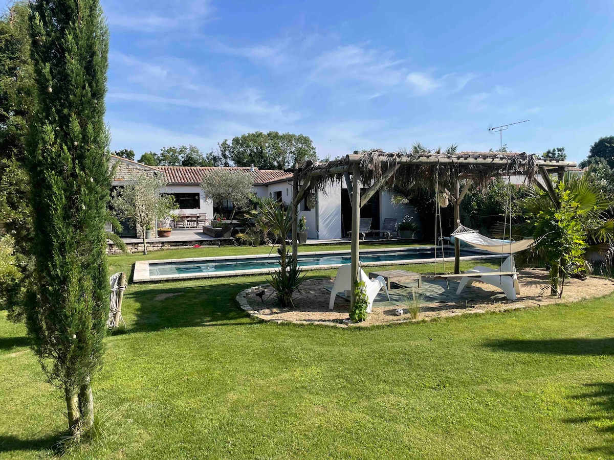 A spacious garden is featured, highlighted by a hammock and seating area under a wooden pergola. In the background, a heated swimming pool is visible, surrounded by lush landscaping and elegant trees, providing a peaceful outdoor setting.
