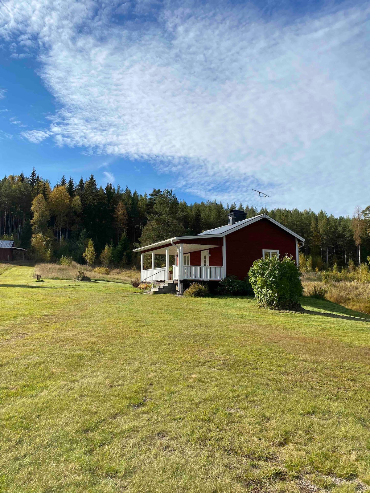 A charming red cottage is positioned on a grassy area, surrounded by forested land. The light blue sky is dotted with white clouds, lending a serene backdrop to the peaceful setting. A spacious front porch is visible, inviting outdoor relaxation.