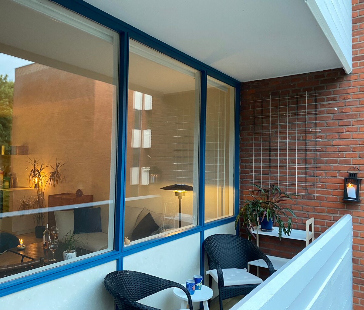 A cozy balcony is displayed, furnished with two black chairs and a small white table. Large windows reveal glimpses of the inviting living space inside, softly lit to create a warm ambiance. Plants are positioned outside, adding a touch of greenery.