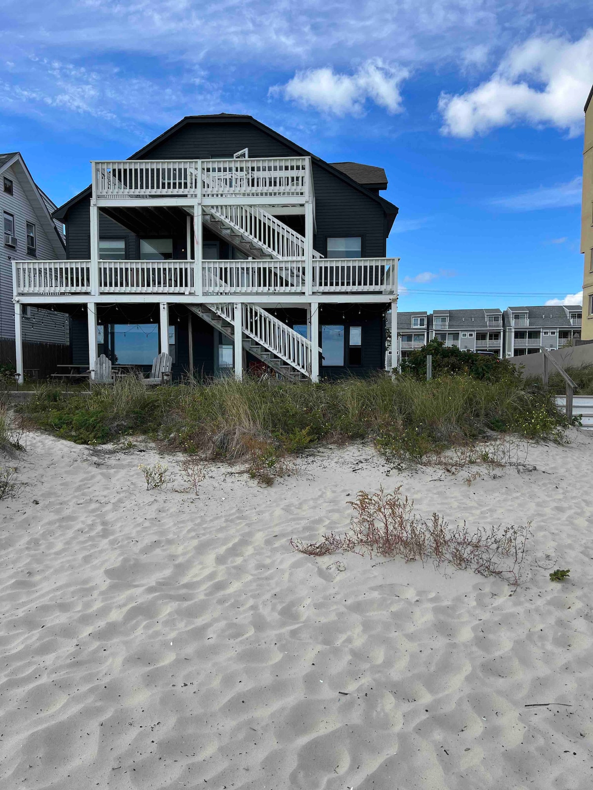 A three-story oceanfront building is depicted, featuring a wraparound deck with multiple steps leading to sandy beach access. The structure is painted in dark tones, set against a backdrop of a clear blue sky and nearby beach houses.