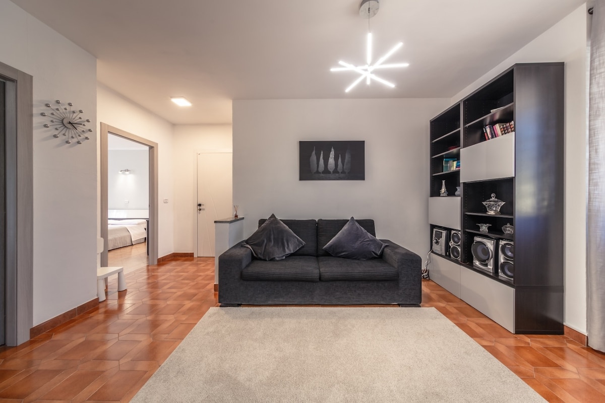 A modern living area is depicted, featuring a dark sofa adorned with decorative pillows. A light fixture with multiple arms illuminates the space, while a plush area rug complements the warm tile flooring. A bookshelf displays various items, adding a personal touch to the room.