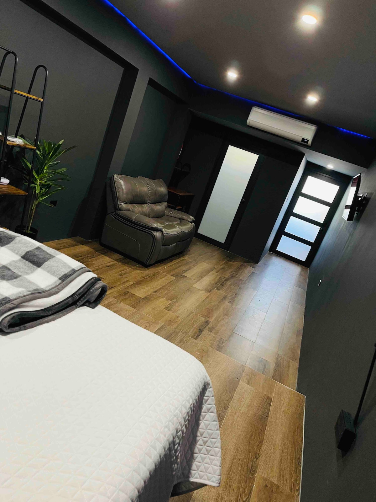 A compact room features a spacious bed covered with a light blanket. A cozy reclining chair is situated against the wall, while sleek wooden flooring extends throughout. Wall-mounted lighting complements the modern design, with a mirrored cabinet door reflecting the room's depth.