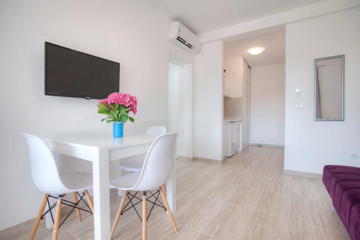 A bright dining area is presented with a white table and four chairs, while a vibrant flower arrangement is displayed in a blue pot. A flat-screen television is mounted on the wall, and a doorway leads towards the kitchen area, showcasing a clean, modern layout.