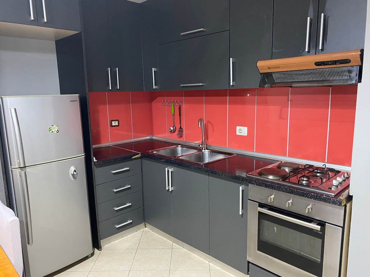 A modern kitchen features sleek gray cabinetry paired with a vibrant red backsplash. Two stainless steel sinks are positioned next to a gas stovetop and oven. A stainless steel refrigerator and organized drawers contribute to a functional cooking space.