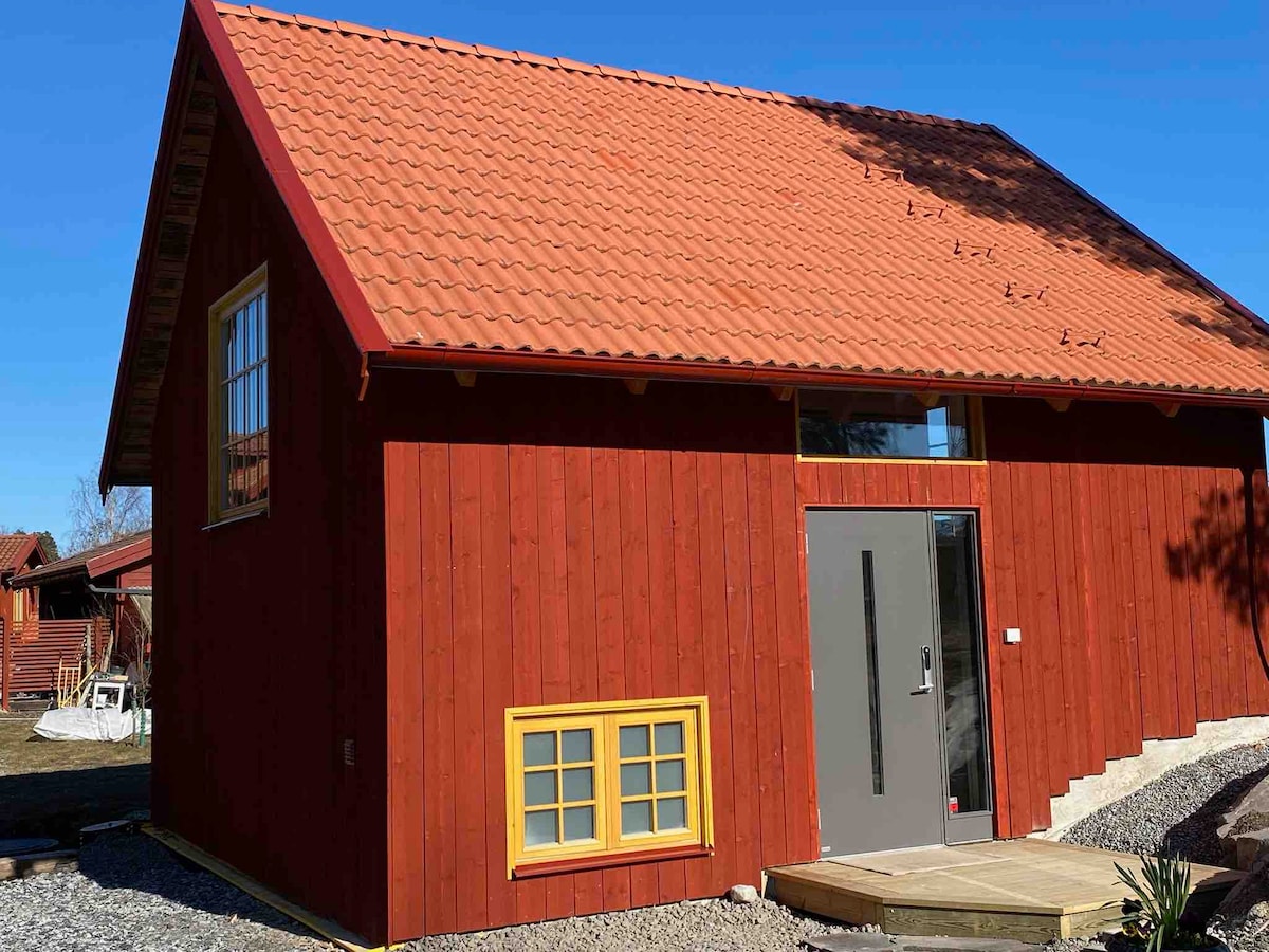 The exterior of a newly constructed red villa is displayed, featuring a distinctive tiled roof and large windows. A modern gray entrance door is highlighted, with a small, bright yellow window in the lower section. A gravel pathway leads to the entrance, surrounded by a serene outdoor area.