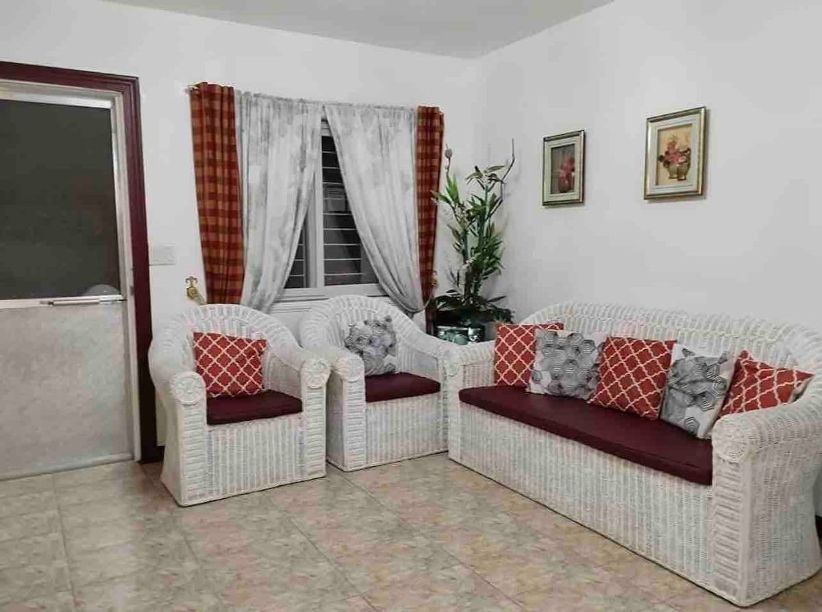 A relaxed seating area features a light-colored rattan sofa with matching armchairs. Decorative red and gray pillows add a touch of color. A window with sheer curtains allows natural light, while framed artwork and a potted plant enhance the overall design.