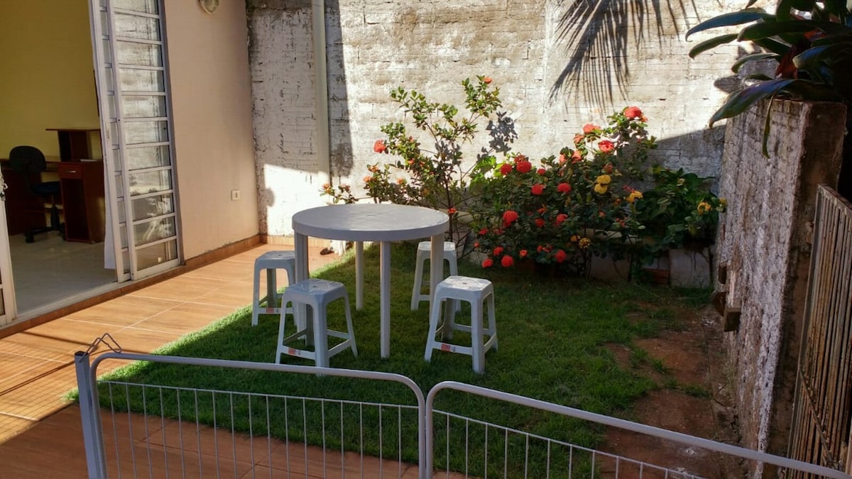 An outdoor space features a small round table surrounded by three stools, all placed on a green lawn. Vibrant flowers in various colors grow around the area, adding a touch of nature. A low fence encloses the space, providing a sense of privacy.