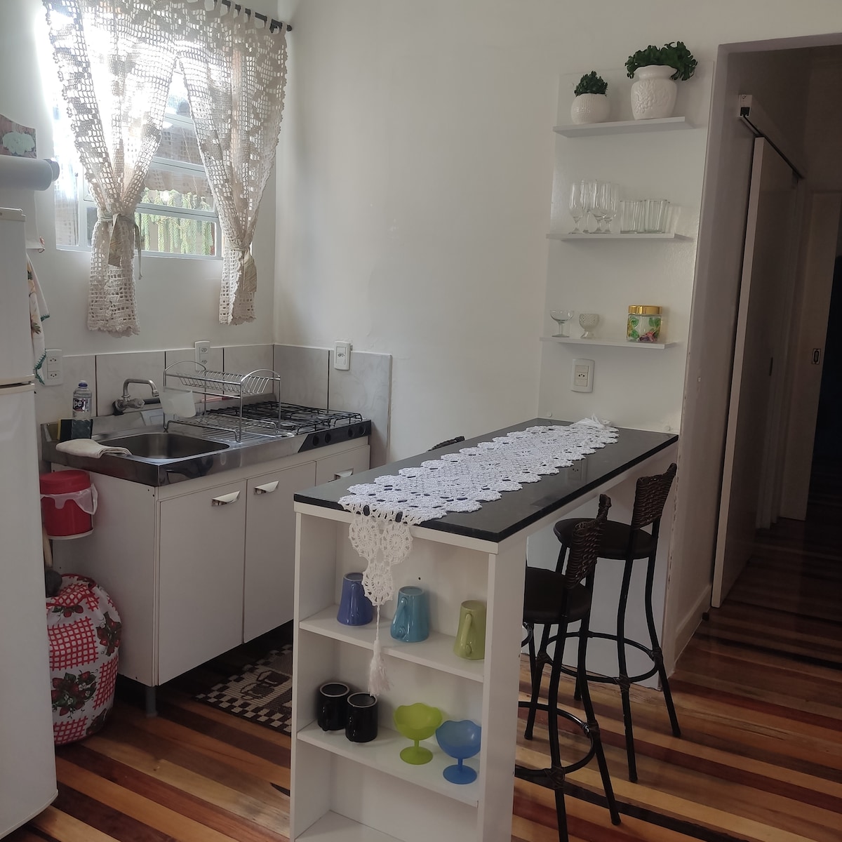 A functional kitchen is highlighted by white cabinetry and a countertop with seating. Natural light filters through a window adorned with lace curtains. Kitchen essentials, including glassware and colorful dishware, are neatly organized on shelves, enhancing the overall accessibility and convenience of the space.