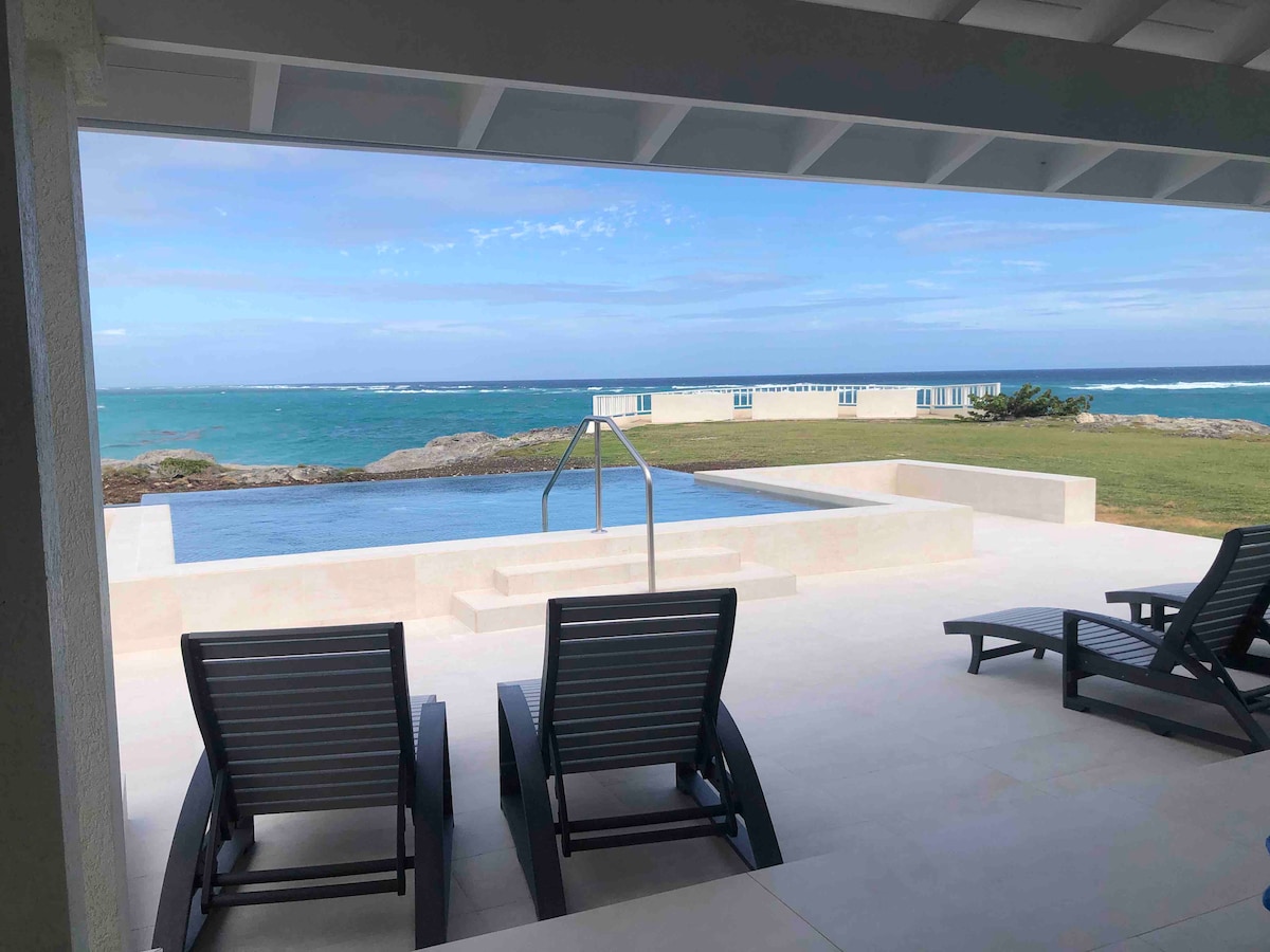 An infinity edge pool extends towards the ocean, framed by a spacious outdoor area. Lounge chairs are arranged on a sunlit patio, with sweeping views of the rugged coastline and clear blue water in the background.