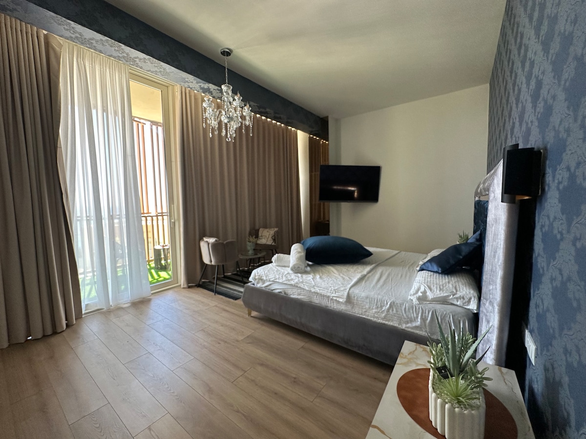 A spacious bedroom is equipped with a large bed featuring plush bedding, complemented by neutral-toned walls. Natural light filters through sheer curtains, enhancing the ambiance. A seating area with a small table is present, along with a mounted television and a decorative plant on a side table.