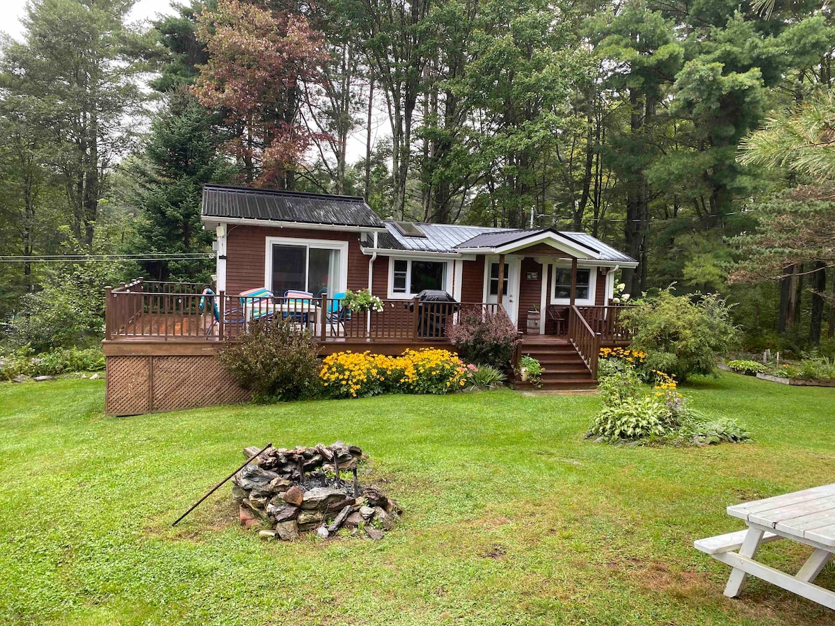 A charming cottage is nestled among trees, featuring a welcoming front deck with outdoor seating. A well-maintained garden with blooming flowers surrounds the property, while a small firepit made of stones is situated on the lush green lawn, providing a serene outdoor space.