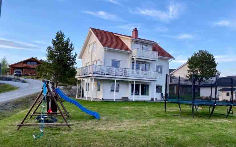 A two-story house is visible, featuring a red roof and multiple balconies. A grassy yard includes a playground with a blue slide and swings. A trampoline stands nearby, with trees providing some shade in the background. The surrounding area is characterized by a quiet residential setting.