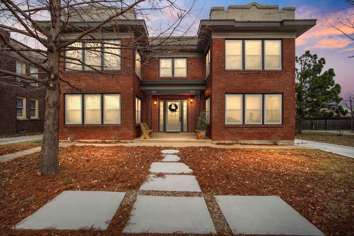 The exterior of the historic condo features a classic brick facade with large windows. A welcoming front porch is flanked by simple wooden chairs. A neatly maintained pathway leads through a naturally landscaped yard with fallen leaves, enhancing the charm of the surrounding area.