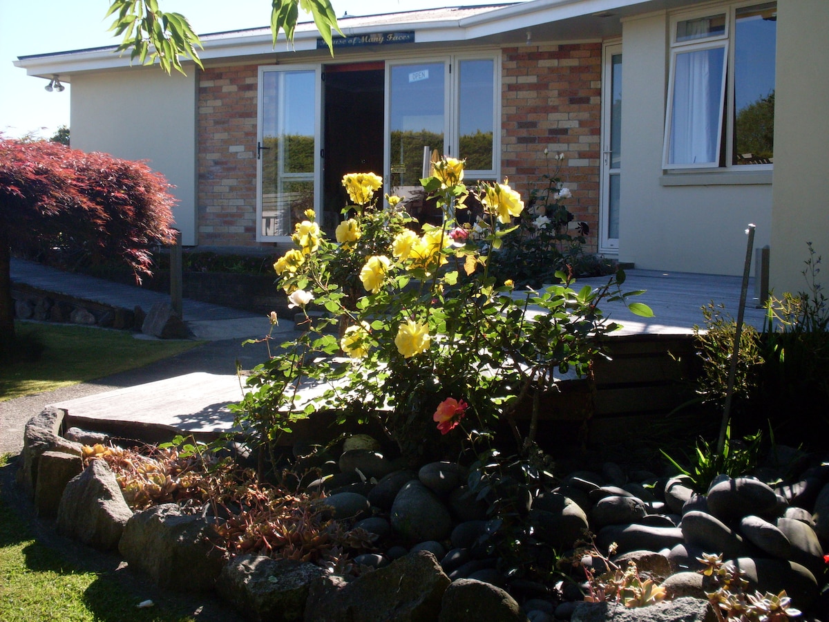 A vibrant flowerbed featuring yellow and orange roses is prominently displayed in the foreground. The well-maintained garden leads to a welcoming brick and wood home, with large windows and a covered porch visible in the background.