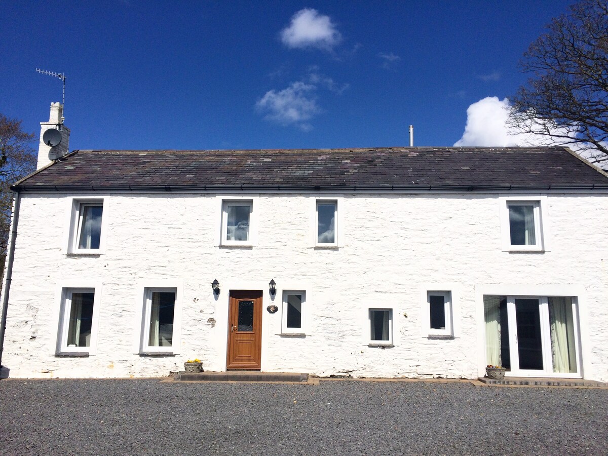 The exterior of a white stone cottage is shown, featuring a central wooden door framed by two outdoor lights. Multiple large windows offer ample natural light. A gravel driveway leads up to the front of the house, surrounded by grassy areas under a clear blue sky.