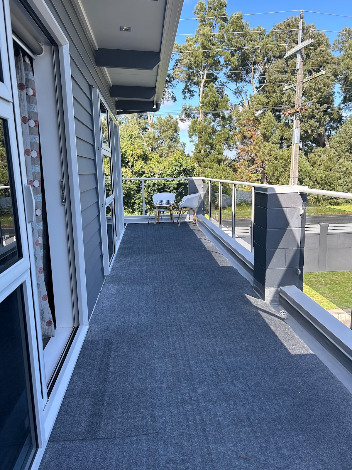 A spacious balcony is presented, featuring a carpeted surface and surrounded by a glass railing. Two white chairs are positioned at the far end, offering views of the greenery beyond the property. Natural light illuminates the area, enhancing the inviting atmosphere.