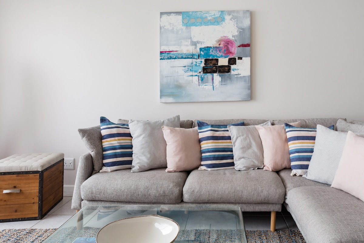A modern living area showcases a spacious gray sectional sofa adorned with decorative pillows in various pastel colors and stripes. A glass coffee table is placed in front, complemented by a textured area rug. An abstract painting hangs on the wall, adding a pop of color.