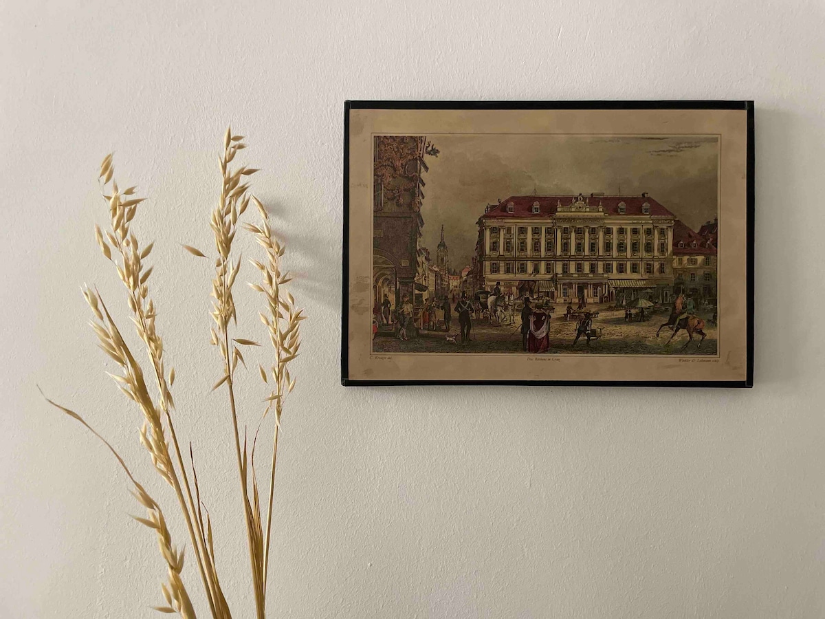 A framed vintage illustration is displayed on a light-colored wall. The artwork depicts a lively street scene with buildings and figures in historical attire. A tall vase with dried grass stands nearby, adding a touch of natural elegance to the setting.