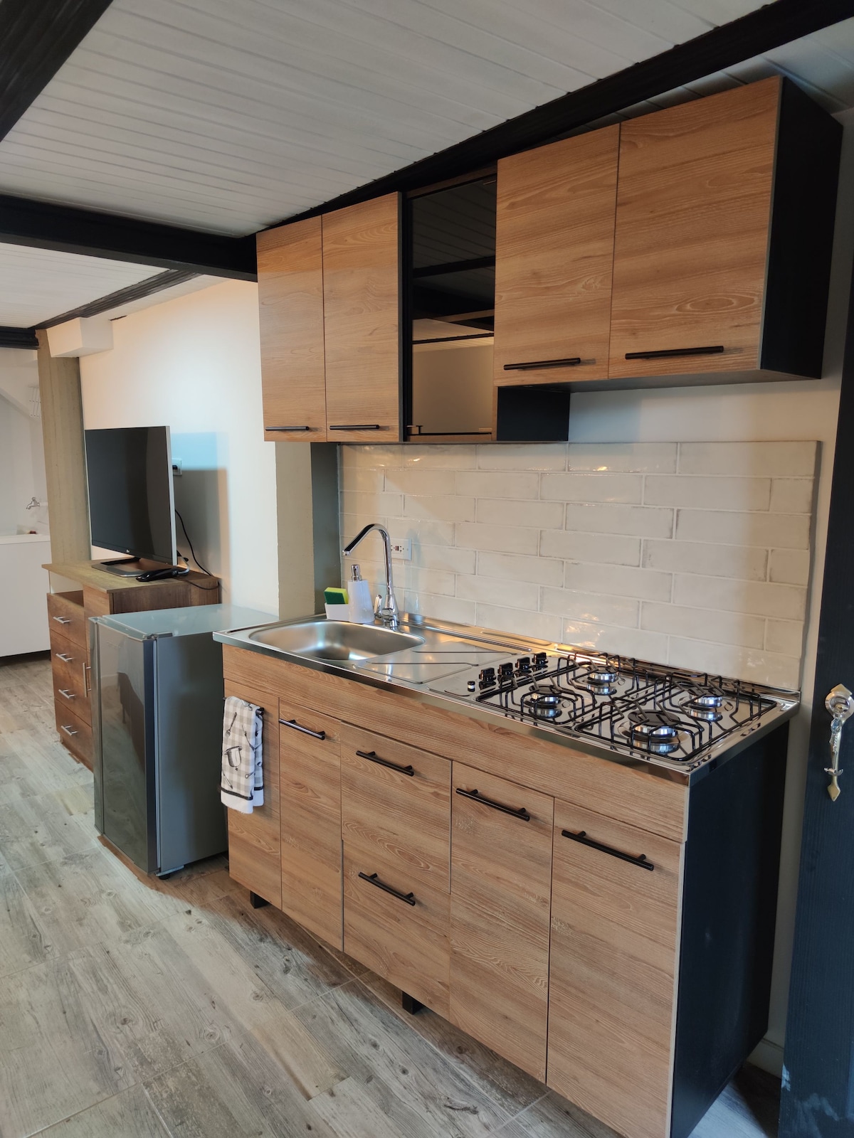 A modern kitchen features wooden cabinets with a minimalist design, complemented by a stainless steel sink and a gas stove. A refrigerator stands to the left, and a small towel hangs neatly by the sink. Natural light illuminates the space, highlighting the clean lines and functionality.