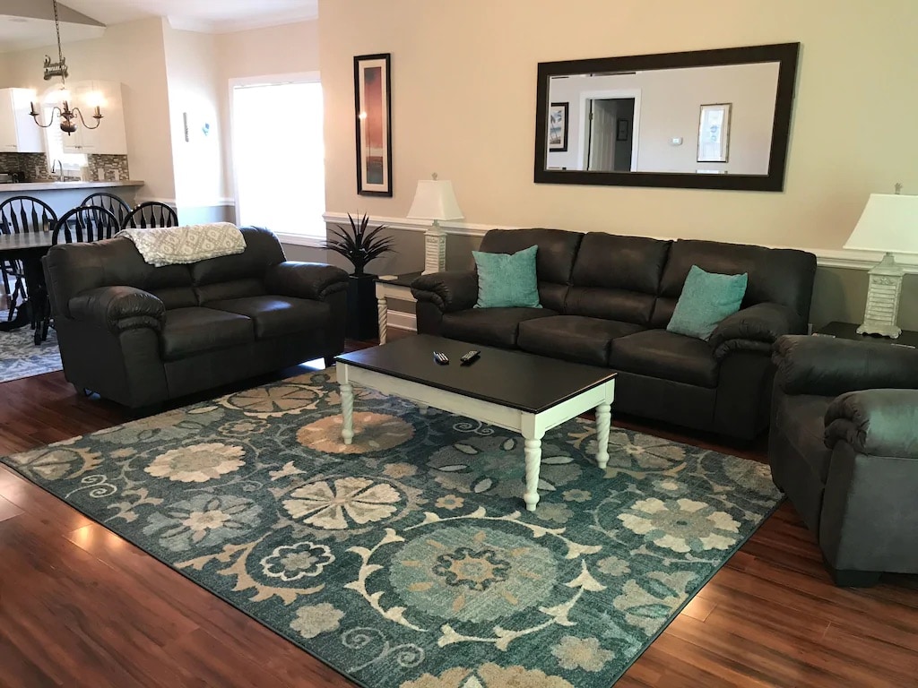 A comfortable living area features two dark sofas and an armchair, arranged around a central coffee table. A decorative area rug with intricate patterns adds warmth to the space, while natural light pours in from a nearby window, creating a welcoming ambiance.