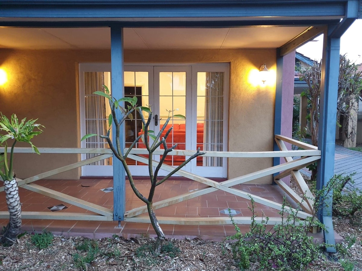 An entrance is highlighted by a sturdy wooden railing and a landscaped area featuring low-lying plants. The exterior walls are painted in warm tones, while double glass doors lead to an interior space illuminated by soft lighting.