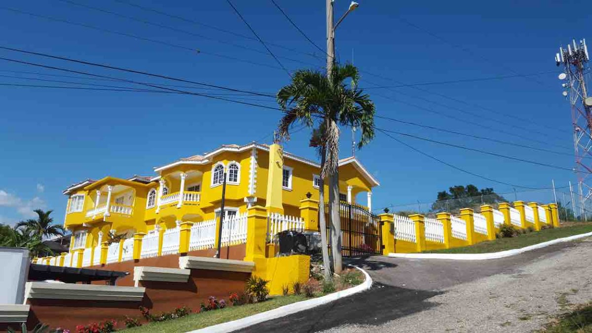 A vibrant yellow property is positioned atop a gentle slope, featuring multiple levels and decorative architectural elements. The surrounding area is landscaped with greenery, and a white fence lines the property. A clear blue sky enhances the overall brightness of the scene.