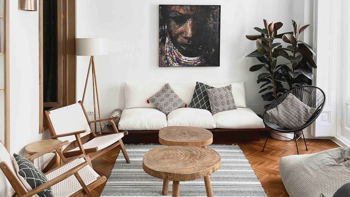 A welcoming living area features a soft sectional sofa adorned with patterned cushions. Natural wood coffee tables sit atop a textured area rug, providing a warm contrast to the polished hardwood floor. A large wall-mounted artwork and potted plants add a touch of character.