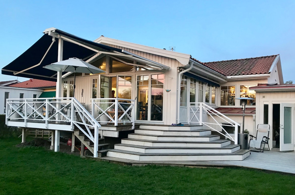 A modern house features a spacious outdoor deck with white railings and steps leading to a well-manicured lawn. Large glass windows allow natural light to fill the interior. An umbrella provides shade over a seating area on the deck, enhancing the outdoor living space.