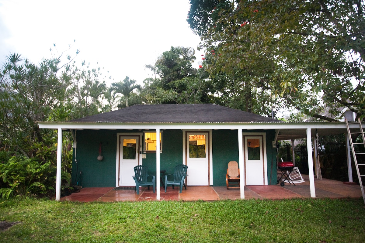 A rustic green cottage with white trim is shown under a canopy of trees. The open lanai features two chairs and a small table, offering a welcoming space for relaxation. The garden surrounds the cottage, creating a peaceful retreat close to nature.